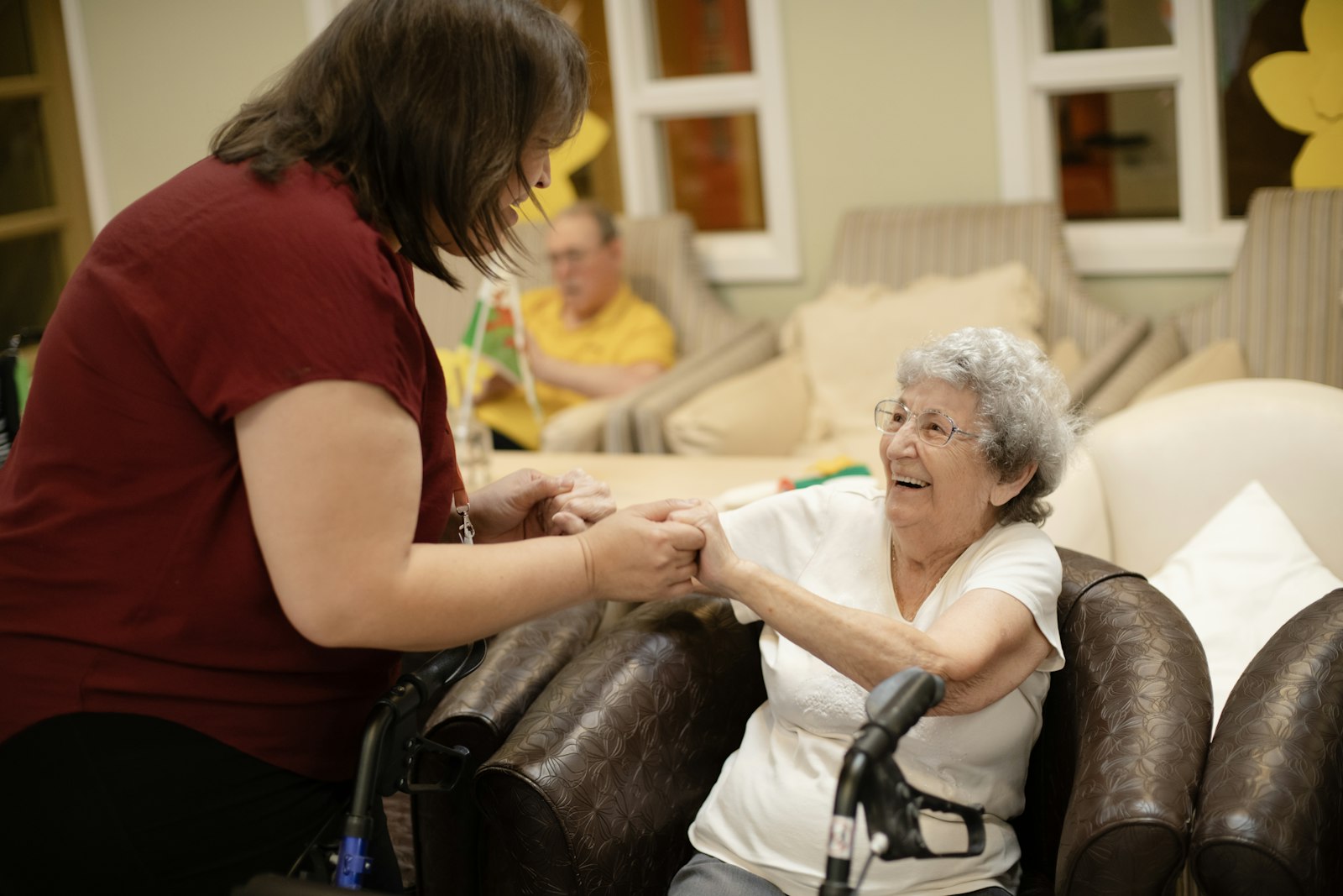 Care assistant holding hands with a smiling elderly woman seated in an armchair